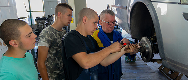 An older male automotive service technician training instructor directing a young male apprentice how to work on automobile brakes while two other apprentices watch.